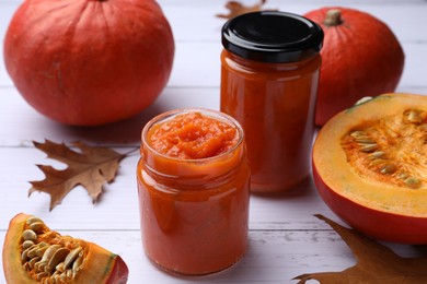 Photo of Jars of delicious pumpkin jam, fresh pumpkin and dry leaves on white wooden table