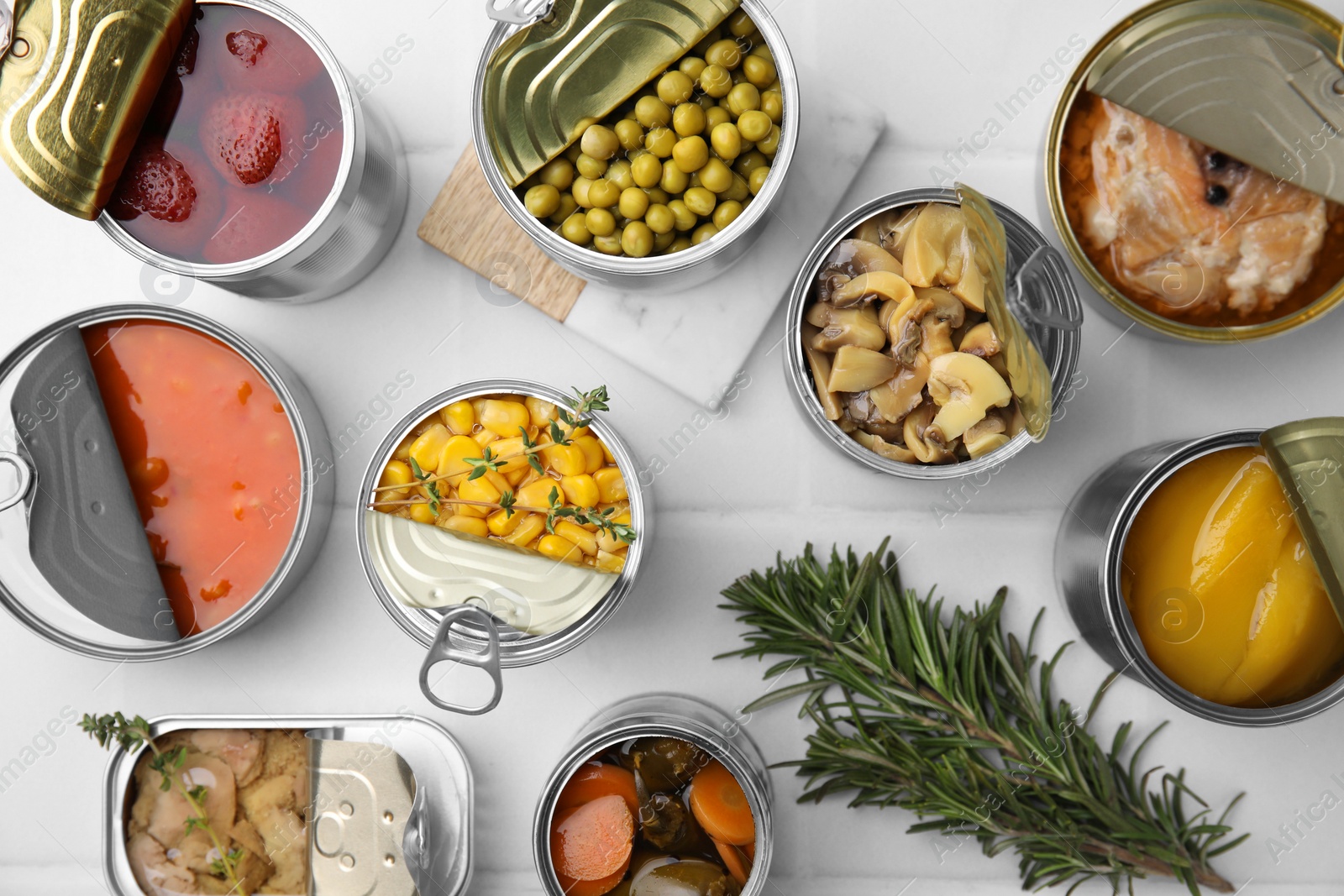 Photo of Open tin cans with different products and rosemary on white tiled table, flat lay