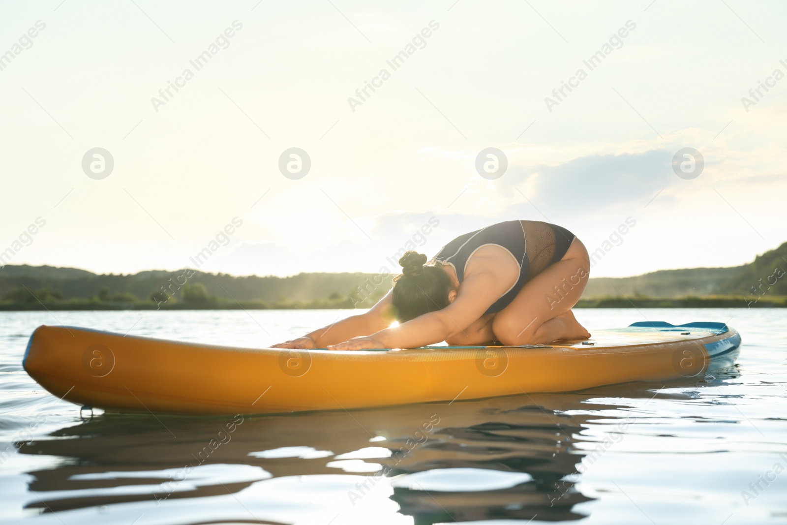 Photo of Young woman practicing yoga on color SUP board on river at sunset