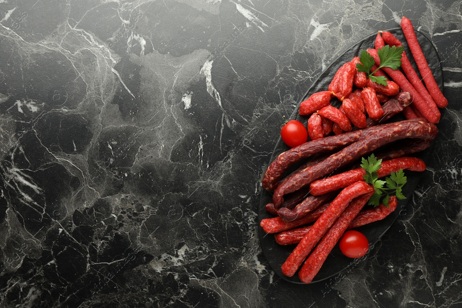 Photo of Thin dry smoked sausages, tomatoes and parsley on black marble table, top view. Space for text