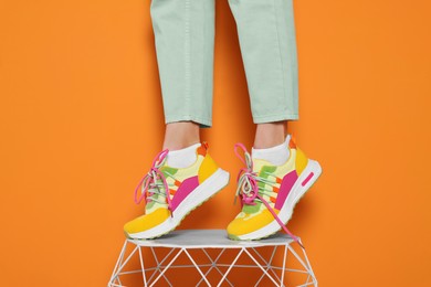 Photo of Woman in stylish colorful sneakers standing on white table against orange background, closeup