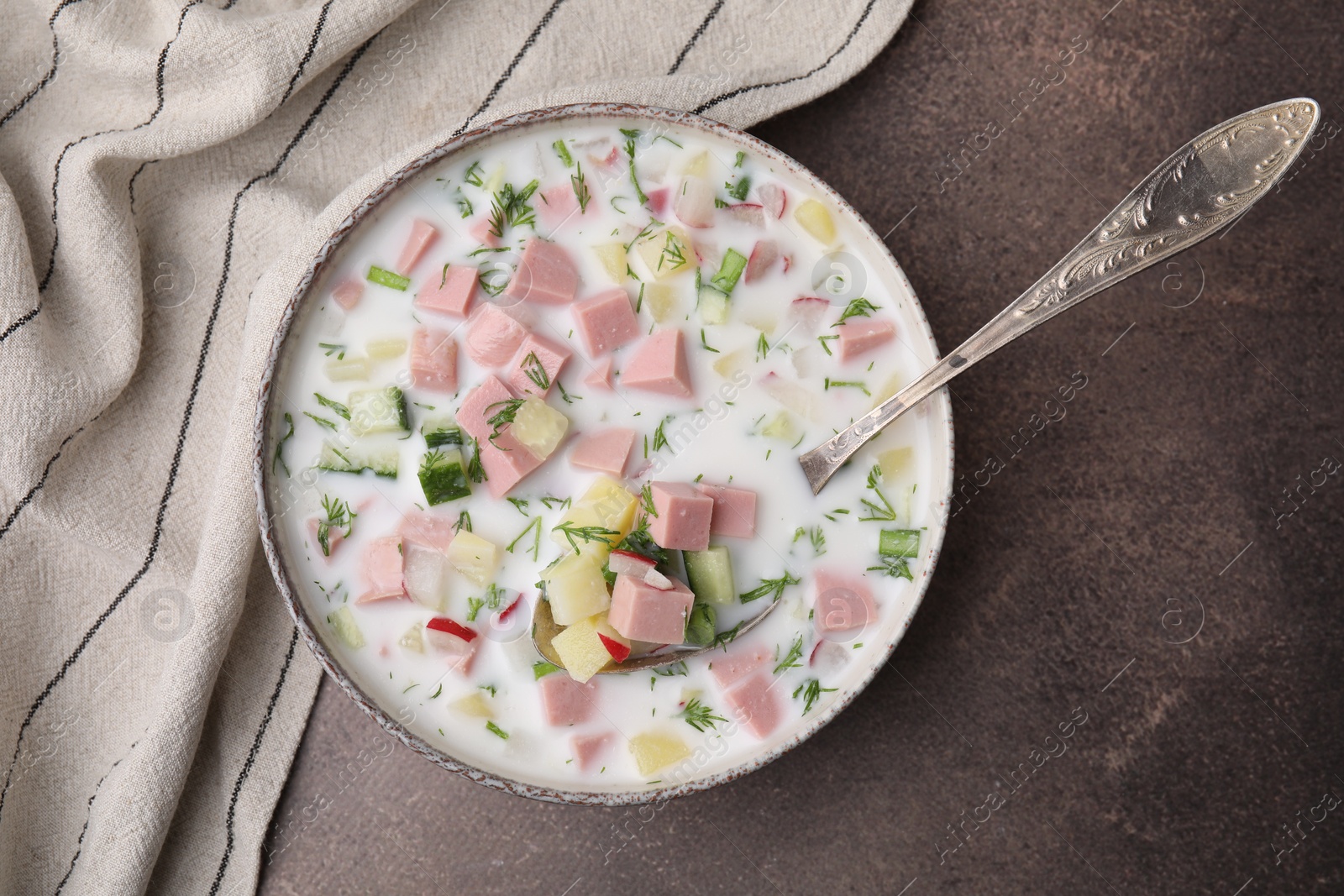 Photo of Delicious cold summer soup (okroshka) with boiled sausage and spoon in bowl on brown table, top view