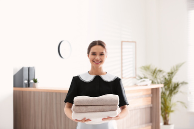 Photo of Young chambermaid holding stack of fresh towels near reception