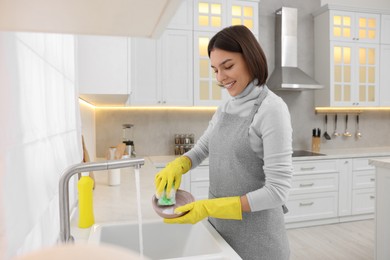 Photo of Happy young woman washing plate above sink in modern kitchen