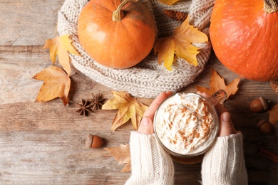 Woman holding cup with pumpkin spice latte on wooden table, flat lay