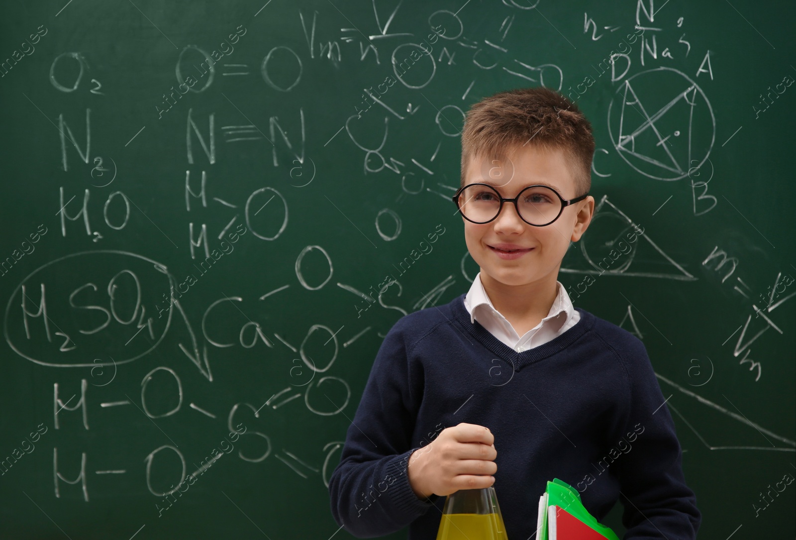 Photo of Schoolboy holding flask near chalkboard with chemical formulas