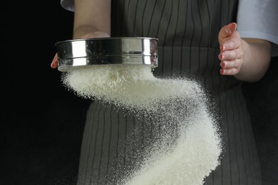 Photo of Woman sieving flour at table against black background, closeup