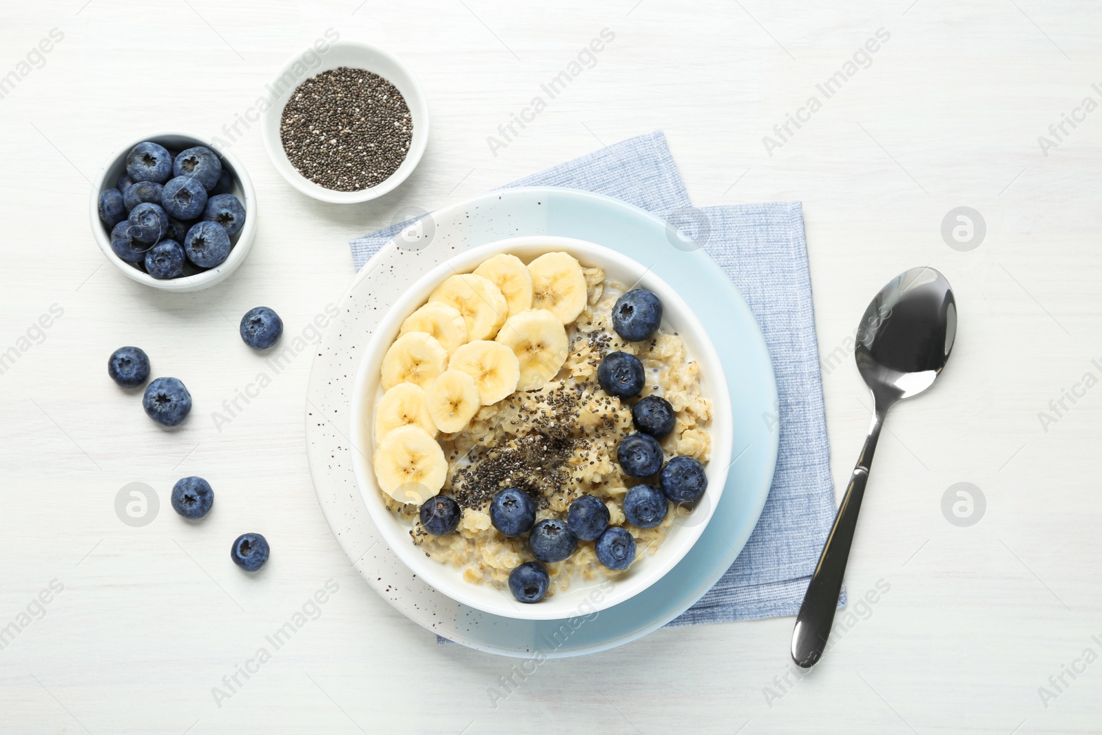 Photo of Tasty oatmeal with banana, blueberries and chia seeds served in bowl on white wooden table, flat lay