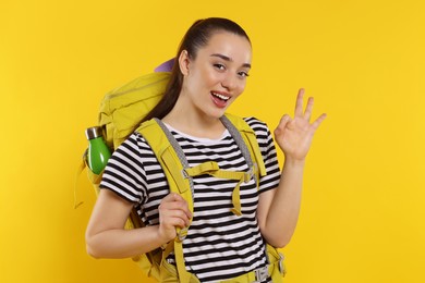 Photo of Smiling young woman with backpack showing OK gesture on orange background. Active tourism