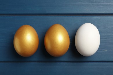 Photo of Golden eggs and ordinary one on blue wooden table, flat lay