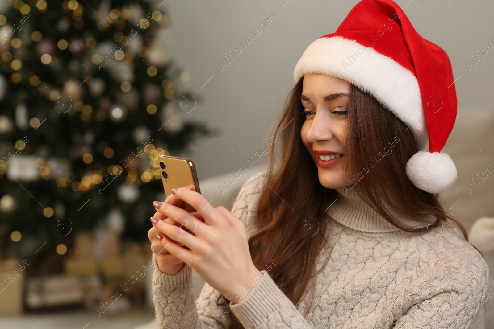 Photo of Beautiful young smiling woman in Santa hat using smartphone near Christmas tree at home, space for text