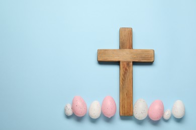 Wooden cross and painted Easter eggs on light blue background, flat lay. Space for text