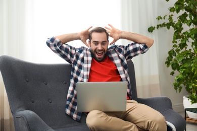 Photo of Emotional young man with laptop celebrating victory on sofa at home