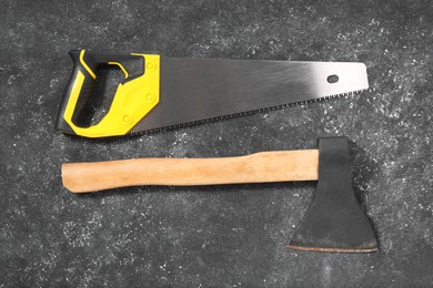 Saw with yellow handle and axe on dark gray textured background, flat lay