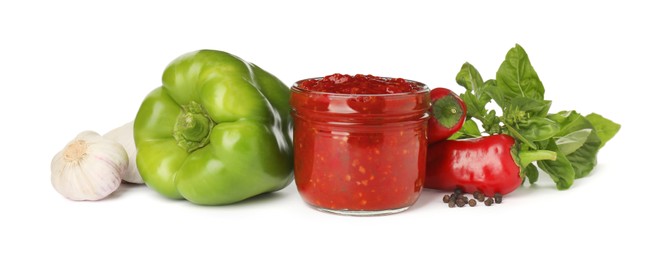 Glass jar of tasty adjika and ingredients on white background