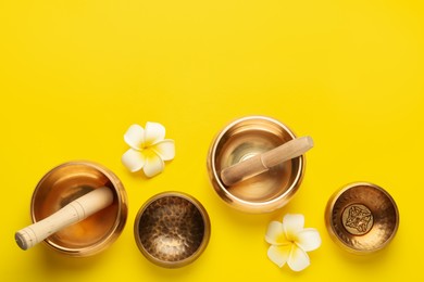 Photo of Golden singing bowls, mallets and flowers on yellow background, flat lay. Space for text