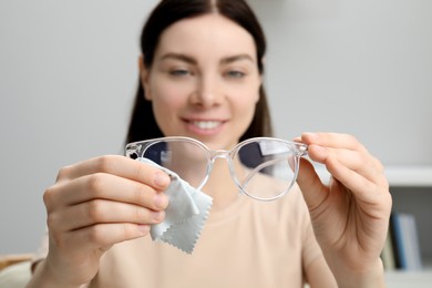 Beautiful woman cleaning glasses with microfiber cloth indoors, selective focus