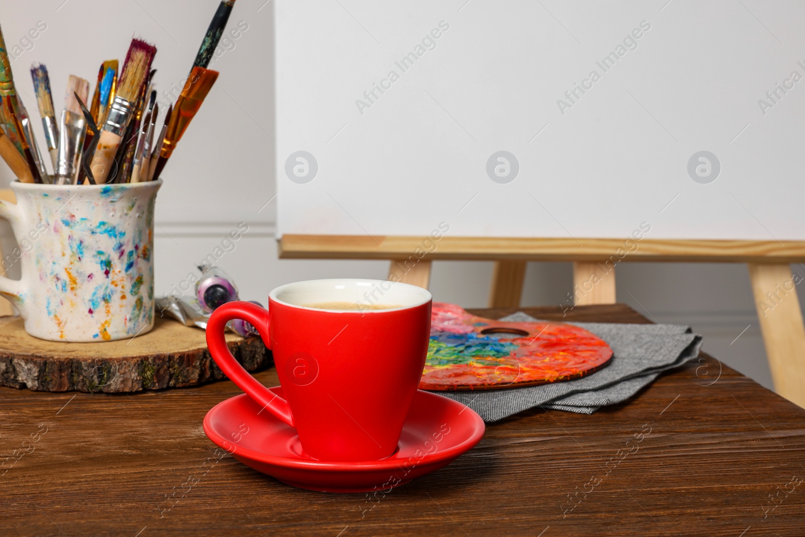 Photo of Easel with blank canvas, cup of drink and different art supplies on wooden table near white wall