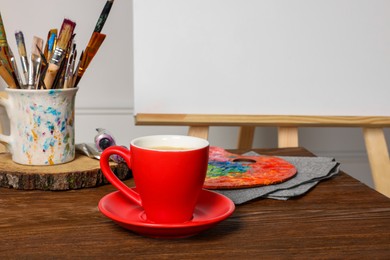 Photo of Easel with blank canvas, cup of drink and different art supplies on wooden table near white wall