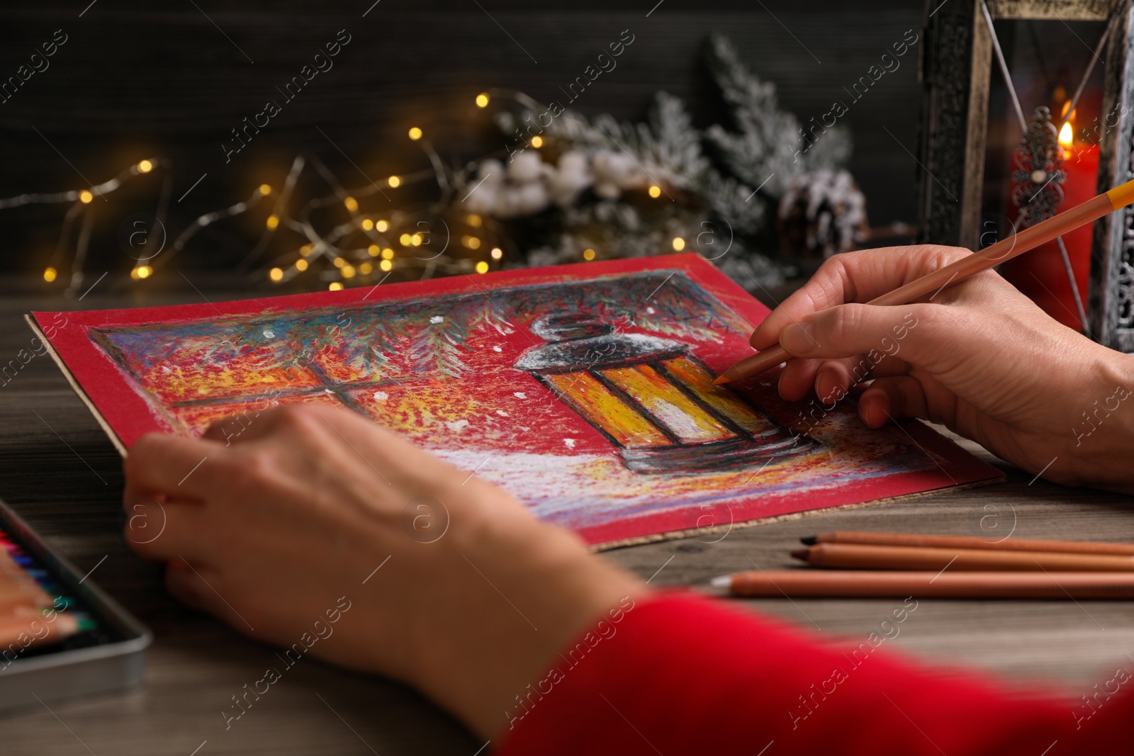 Photo of Woman drawing beautiful lantern with pastel pencil at wooden table against blurred lights, closeup