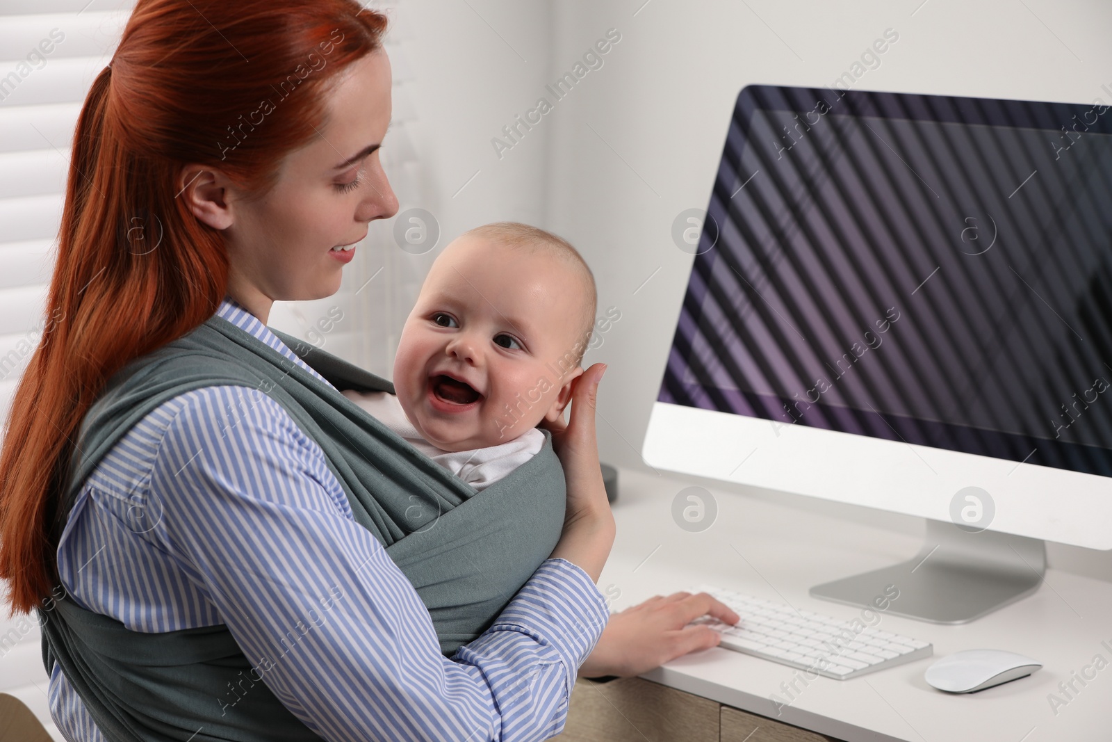 Photo of Mother holding her child in sling (baby carrier) while using computer at workplace