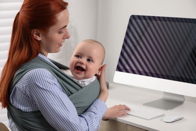Mother holding her child in sling (baby carrier) while using computer at workplace