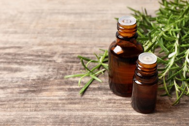 Bottles of essential oil and fresh rosemary sprigs on wooden table, space for text