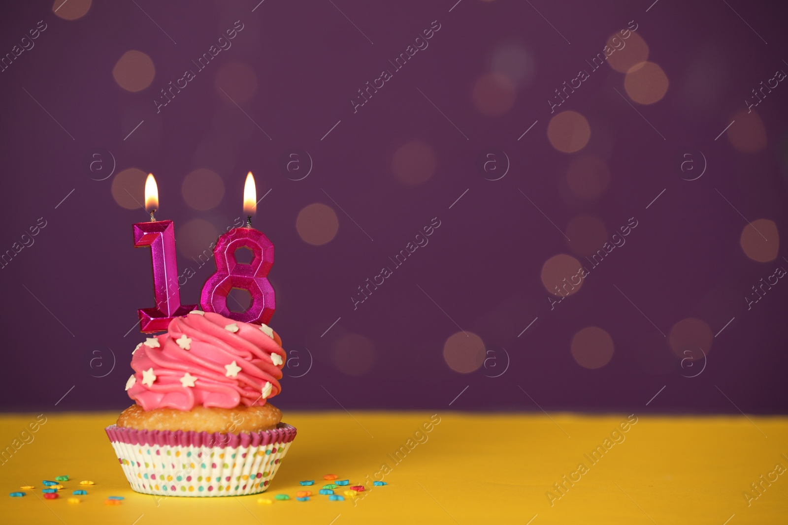 Photo of Coming of age party - 18th birthday. Delicious cupcake with number shaped candles on yellow table against blurred lights, space for text