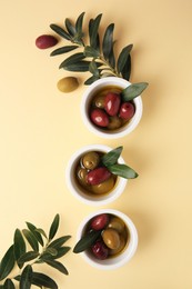 Bowls with different ripe olives and leaves on beige background, flat lay