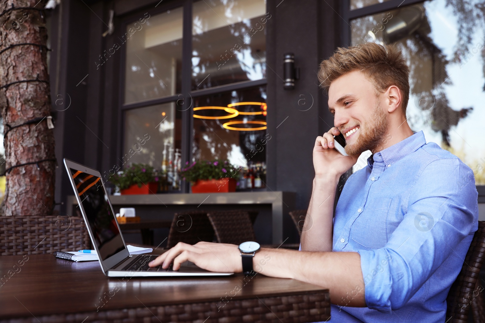 Image of Happy young man talking on phone while using laptop at outdoor cafe