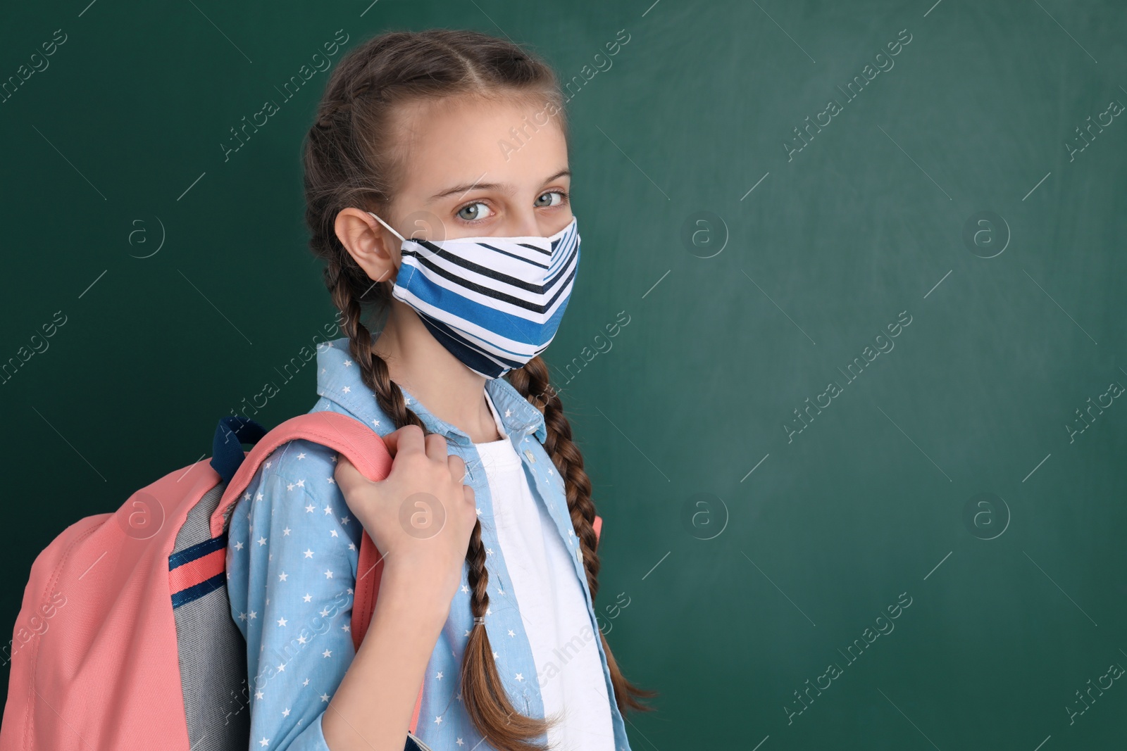 Photo of Schoolgirl wearing protective mask and backpack near green chalkboard, space for text. Child's safety from virus
