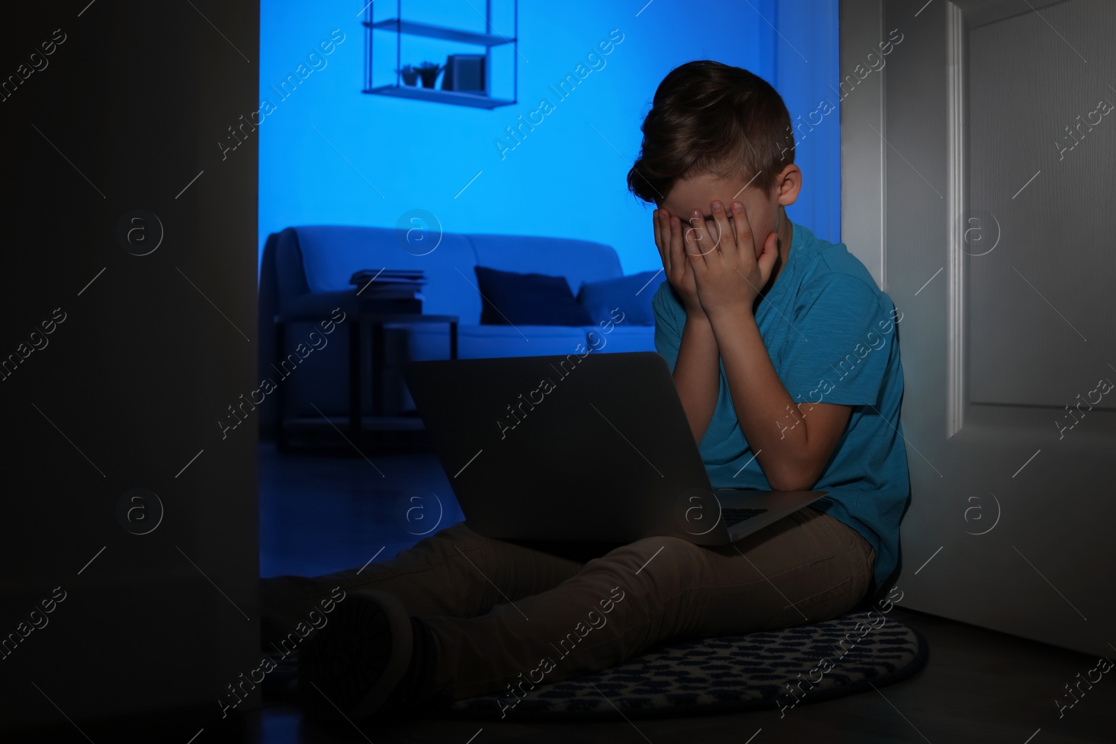 Photo of Frightened little child with laptop on floor in dark room. Danger of internet