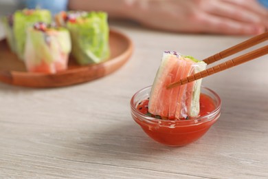 Woman dipping delicious spring roll wrapped in rice paper into sauce at white wooden table, closeup