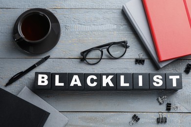 Photo of Black cubes with word Blacklist, cup of coffee and office stationery on grey wooden table, flat lay