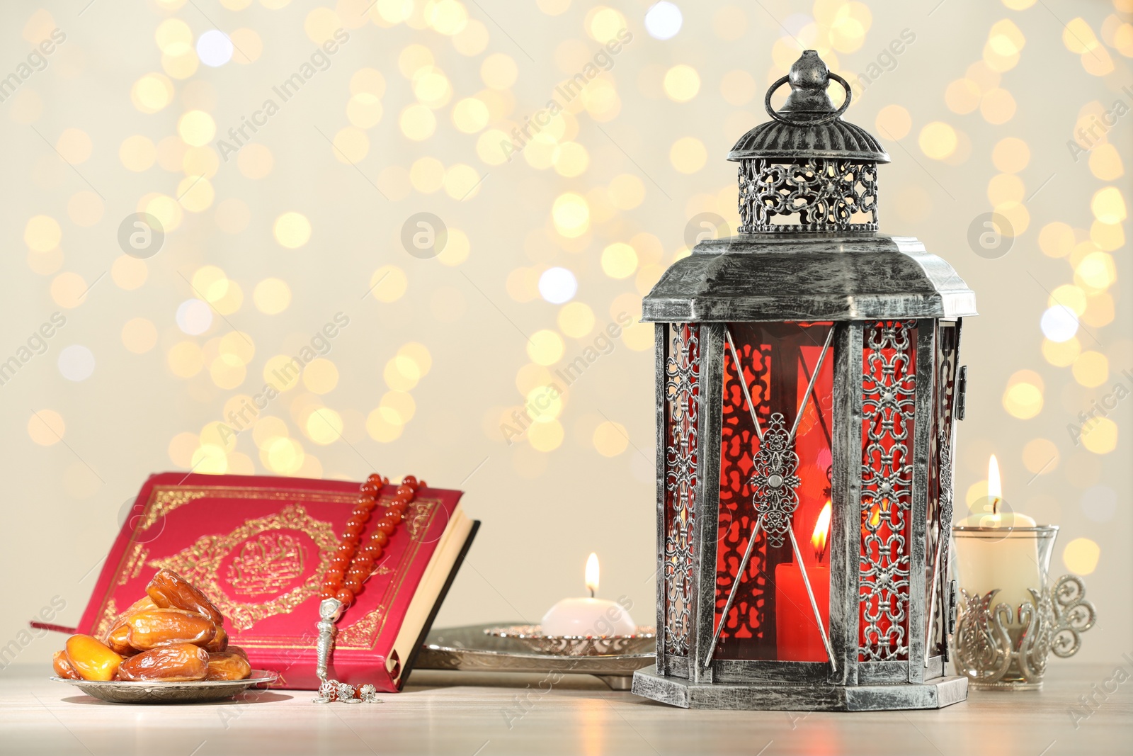 Photo of Arabic lantern, Quran, misbaha, candles and dates on table against blurred lights