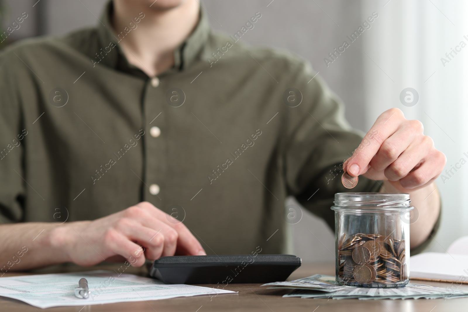 Photo of Financial savings. Man putting coin into glass jar while using calculator at wooden table, closeup