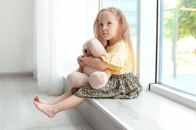 Photo of Pretty little girl with teddy bear near window in room