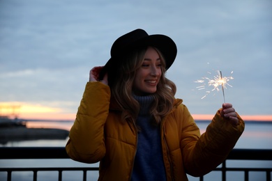 Photo of Woman in warm clothes holding burning sparkler near river
