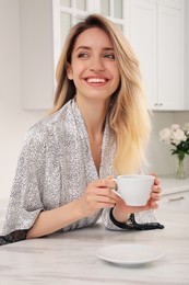 Pretty young woman in beautiful silk robe with cup of coffee at kitchen table
