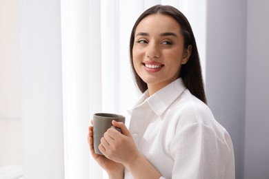 Beautiful woman with cup of drink near window at home, space for text. Lazy morning