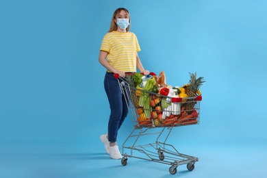 Young woman in medical mask with shopping cart full of groceries on light blue background