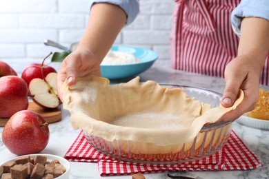 Woman putting dough for apple pie into baking dish at white marble table, closeup