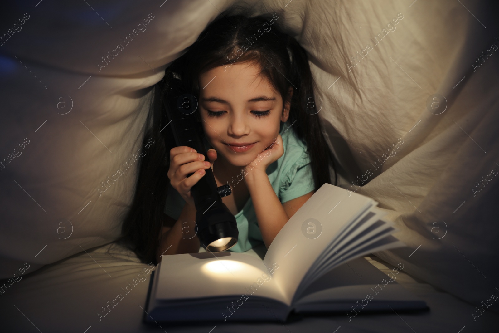 Photo of Little girl reading fairy tale under blanket in dark bedroom