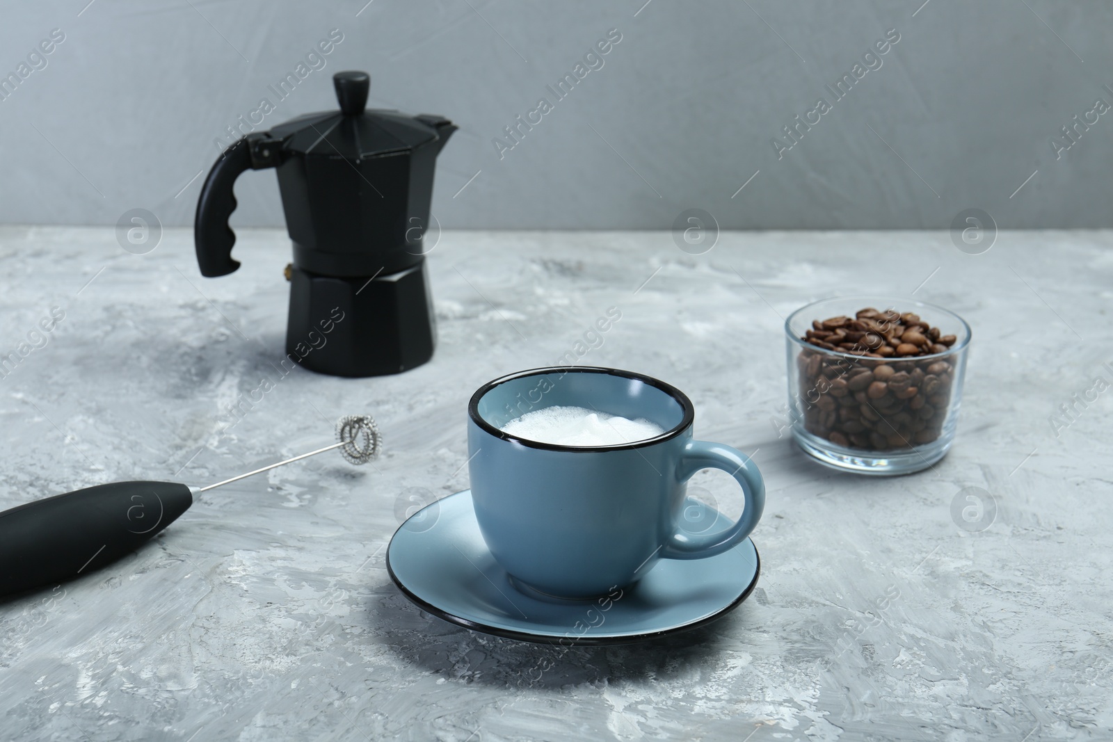 Photo of Mini mixer (milk frother), whipped milk in cup and coffee beans on grey textured table