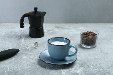 Photo of Mini mixer (milk frother), whipped milk in cup and coffee beans on grey textured table