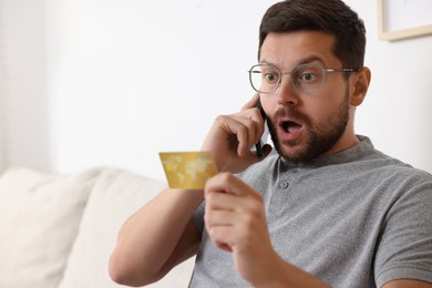 Emotional man with credit card talking on smartphone indoors, space for text. Be careful - fraud