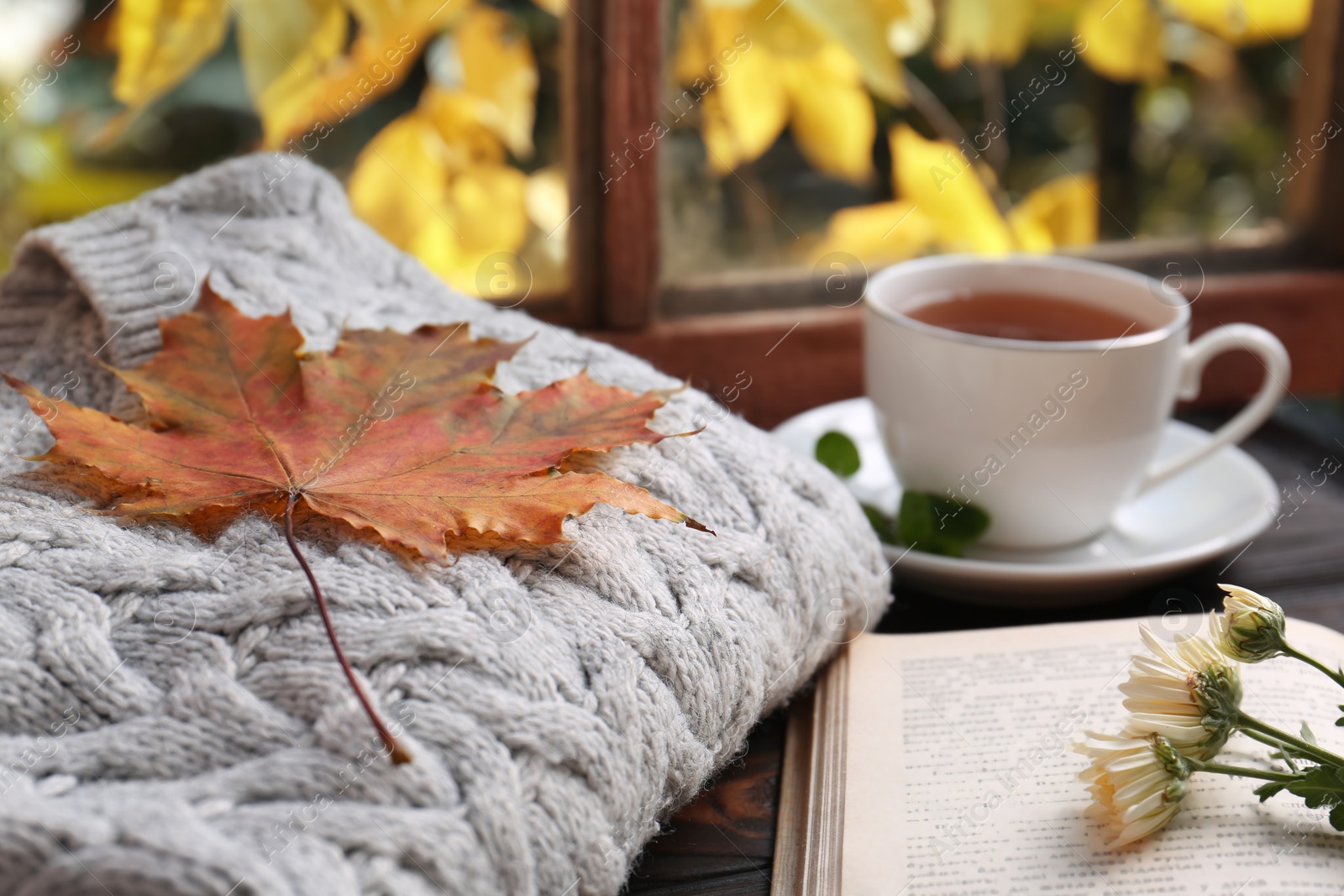 Photo of Cup of aromatic tea, soft sweater and book on wooden windowsill indoors, closeup. Autumn atmosphere