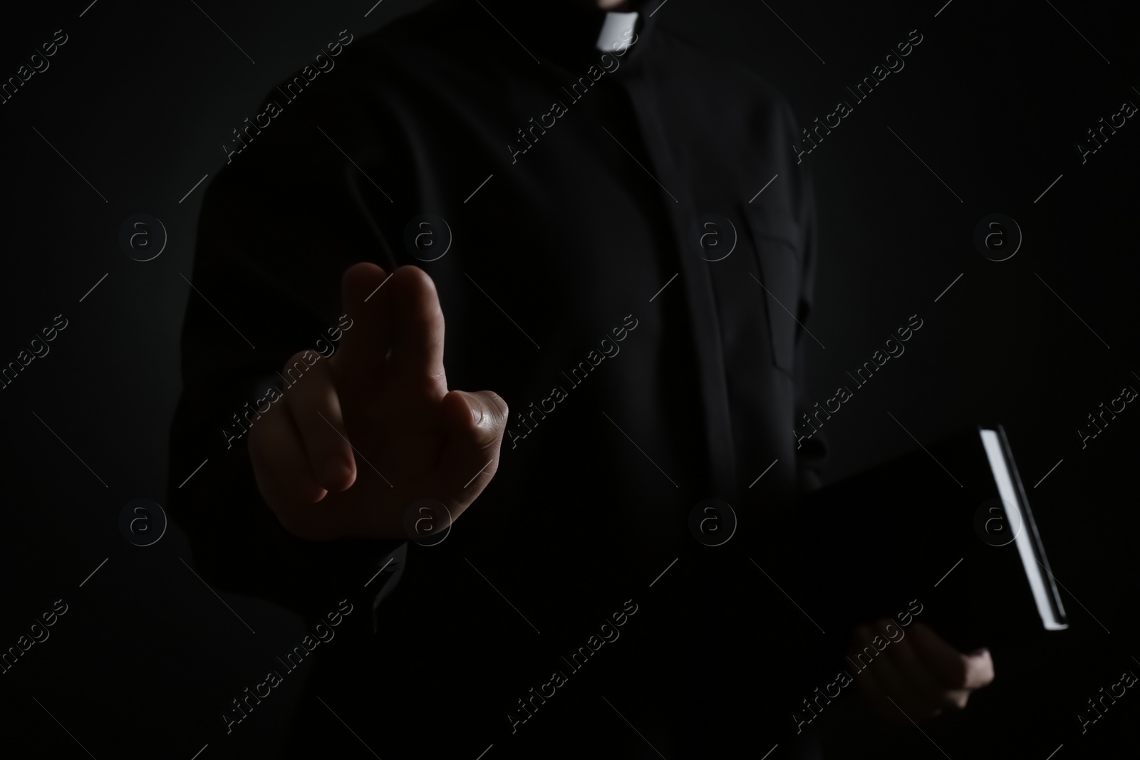 Photo of Priest with Bible making blessing gesture on dark background, closeup
