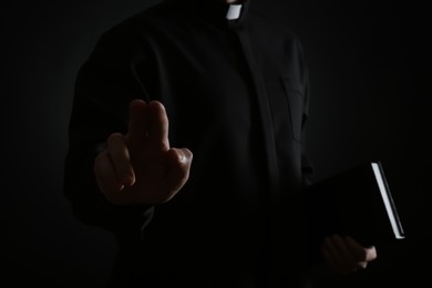 Photo of Priest with Bible making blessing gesture on dark background, closeup