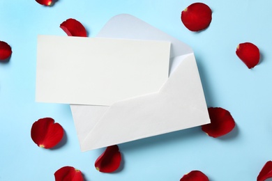 Photo of Blank greeting card, envelope and rose petals on light blue background, flat lay. Valentine's day celebration